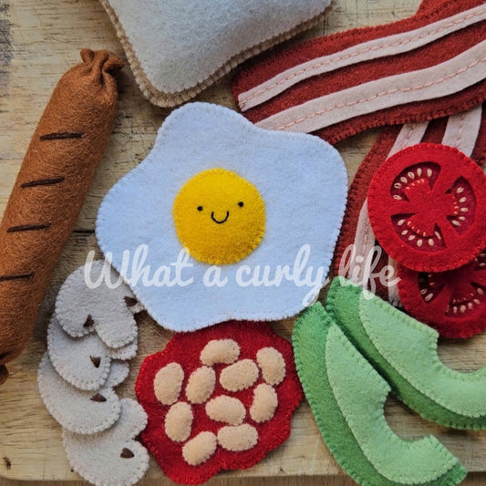 Felt food items including an egg, bread, tomatoes, and beans on a wooden surface with 'What a curly life' text.